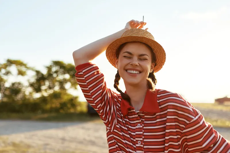 Smiling woman with braids wearing a straw hat outdoor. Lifestyle portrait in summer sunlight. Positive lady feels happy and free during vacation. Golden hour lighting creates warm glow.
