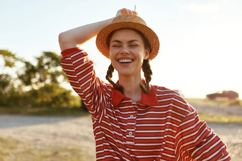 Young woman in a straw hat and striped shirt laughs outdoors while enjoying a sunny day. Her joyful expression creates an authentic lifestyle scene captured in warm golden sunlight.