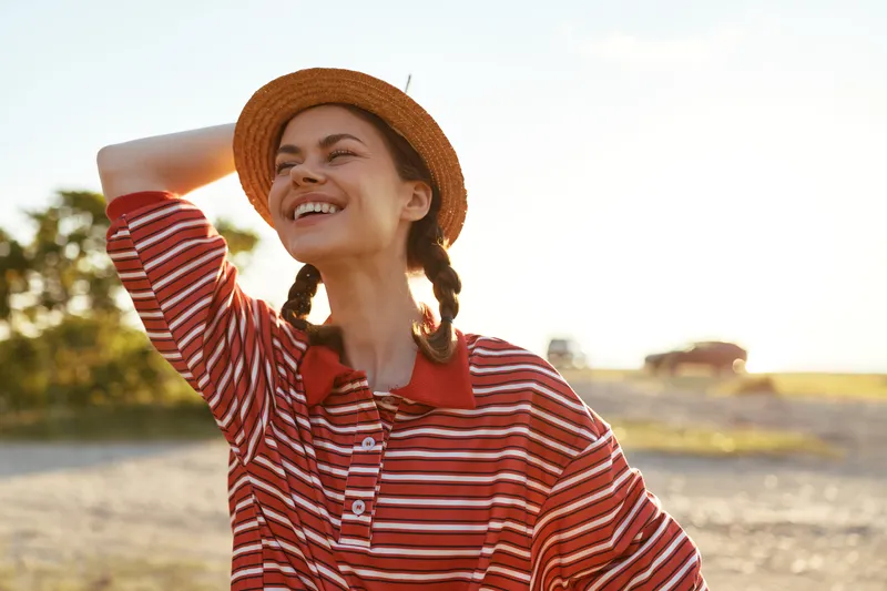 A happy young woman in a striped shirt and a stylish straw hat enjoys a sunny day outdoors. Lifestyle concept for summer vacation, freedom, joy, and travel in a beautiful natural landscape.
