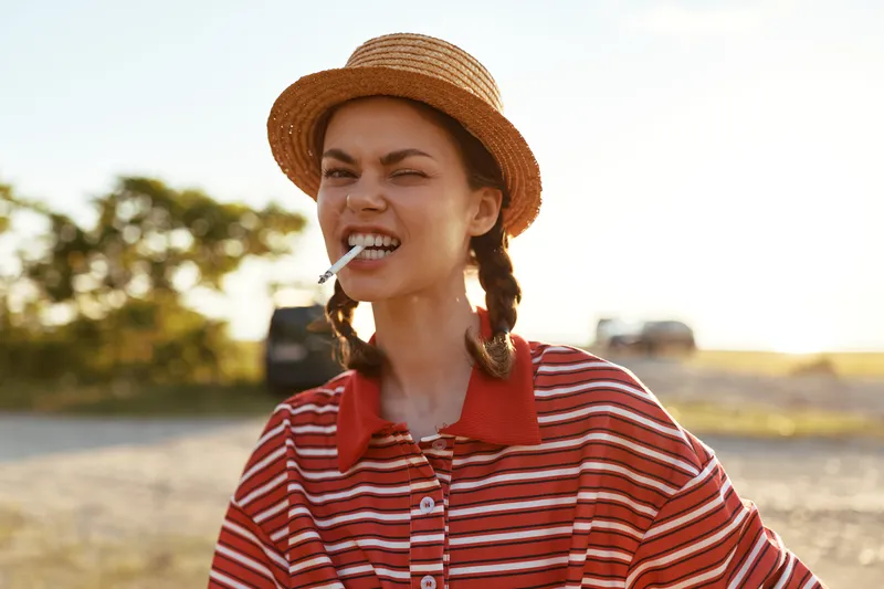 Young woman in a straw hat and striped shirt winks while holding a cigarette in her mouth outdoors. Authentic lifestyle portrait of a girl with braids enjoying a sunny day in the countryside.