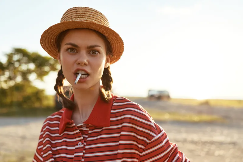 Young woman in straw hat and striped polo shirt holding cigarette in mouth posing outdoors. Concept of rebellious lifestyle and authentic portrait of a lady in rural setting.
