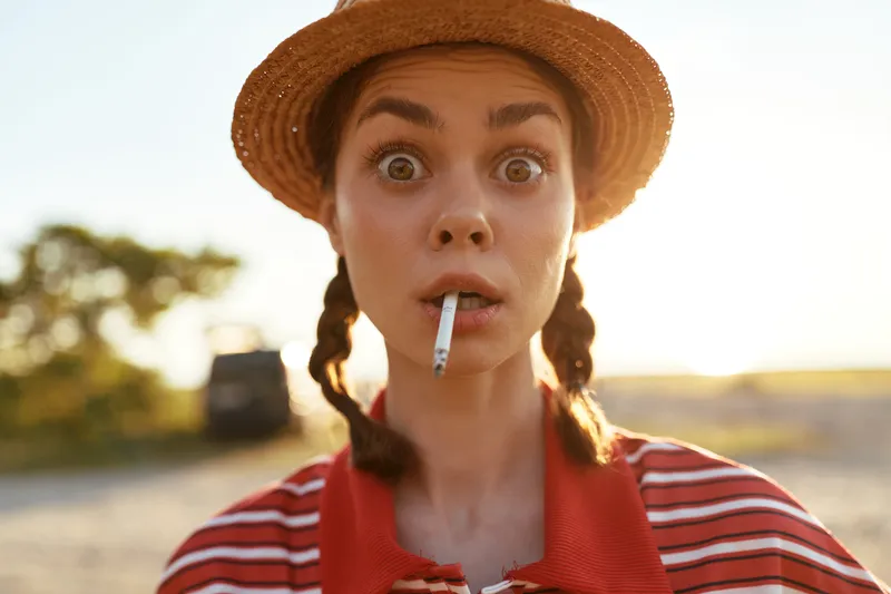 Surprised woman lifestyle portrait with a cigarette in her mouth, wearing a sun hat. Outdoor summer vibe, natural light, emotional expression, creative photography, candid shot.
