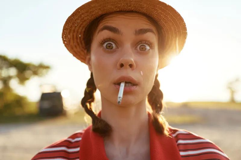 Surprised woman lifestyle portrait outdoors wearing a straw hat and striped shirt with a cigarette in her mouth. Golden hour sun flare lighting concept for urban youth style.