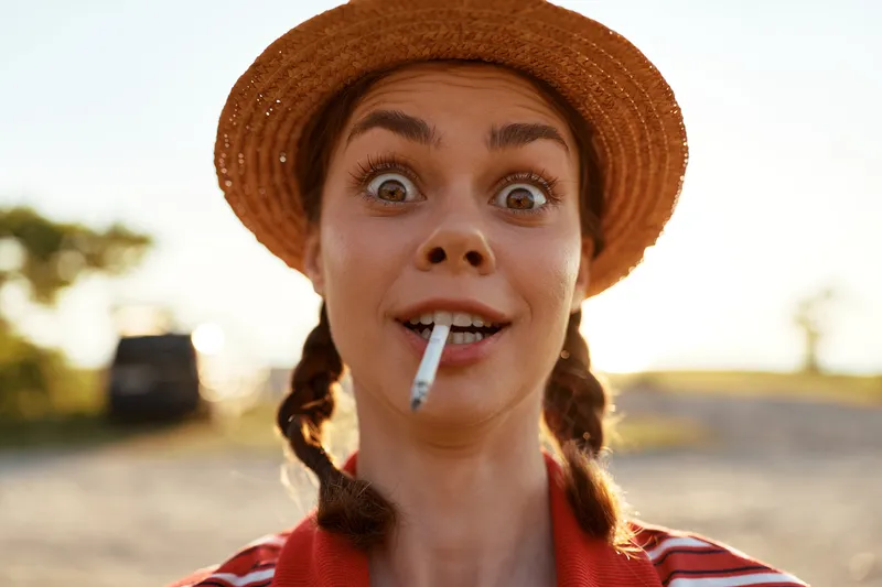Young woman with braided hair wearing a straw hat holds a cigarette in her mouth. Lifestyle portrait outdoors in soft sunlight. Concept of rebel mood and freedom.