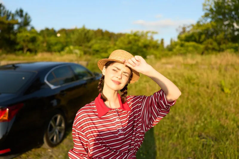 A young woman wearing a straw hat and striped shirt stands outside near a car. Sunny lifestyle portrait in a field with golden hour light. Concept of summer travel and leisure.