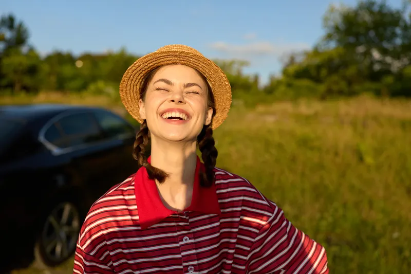 Cheerful young woman with braids in a straw hat laughing outdoors in a meadow at sunset. The happy girl enjoys a sunny summer lifestyle near a car in the field.