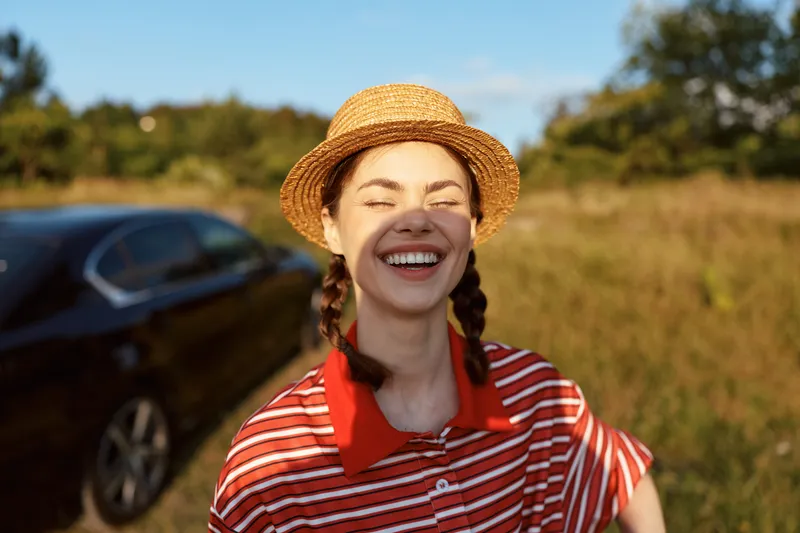 A happy young woman lifestyle portrait outdoors in a straw hat. She laughs in the sun near a car. Concept for freedom, joy, summer travel, and leisure vacation.
