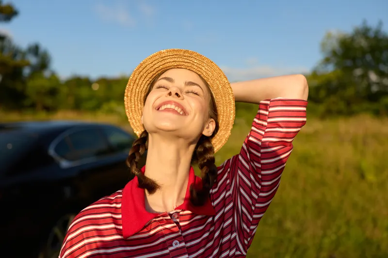 Happy woman enjoying lifestyle in summer nature. Smiling girl wearing a straw hat posing outdoors with closed eyes. Scenic landscape view, golden hour sunlight, freedom and relaxation concept.