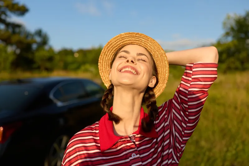 Smiling young woman in a straw hat and striped shirt enjoys the warm sunlight in a green field near a black car during a summer road trip, feeling happy and carefree.