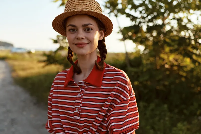 Smiling woman, lifestyle portrait outdoors, wearing straw hat and striped shirt. The young female enjoys peaceful summer evening, natural sunlight, glowing skin, authentic rural scenery.