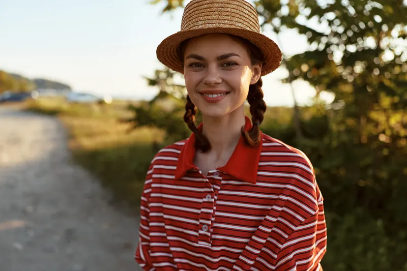 Smiling woman lifestyle portrait posing in a straw hat and a striped shirt outdoors during summer evening sunset. Concept for travel, leisure, vacation, happiness, joy, and female beauty.
