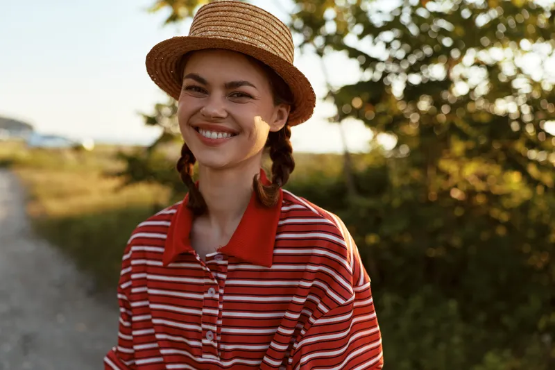 Smiling young woman in a straw hat and striped shirt enjoys a sunlit day outdoors, capturing an authentic lifestyle moment of happiness and relaxation in the warm golden evening light.