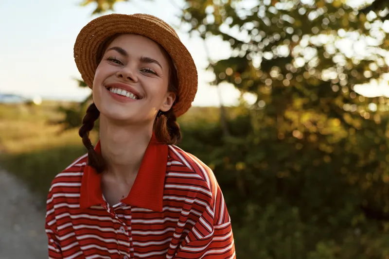 Smiling woman wearing straw hat and striped shirt outdoors in summer. Beautiful lady enjoying lifestyle portrait with natural sunlight, joyful expression and casual clothing style.