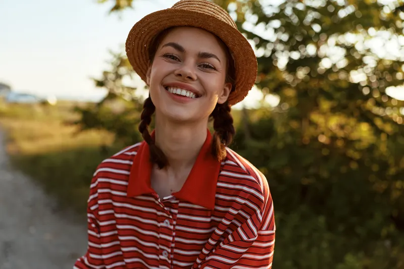 Smiling young woman in straw hat and striped shirt enjoys sunny summer day in nature. Portrait of a happy lady with braided hair laughing during her authentic casual lifestyle walk.