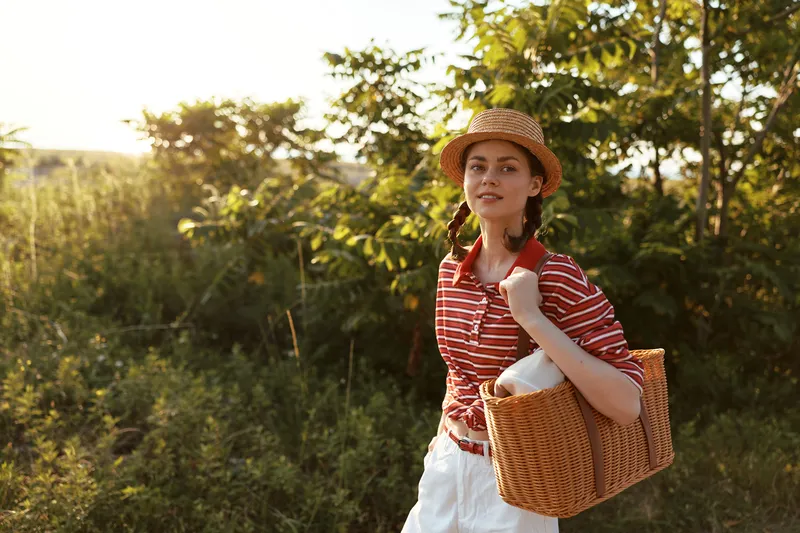 Young woman with straw hat and basket walking in sunny park. Casual lifestyle portrait outdoors. Natural golden hour lighting creates a warm atmosphere for holiday and travel concept.