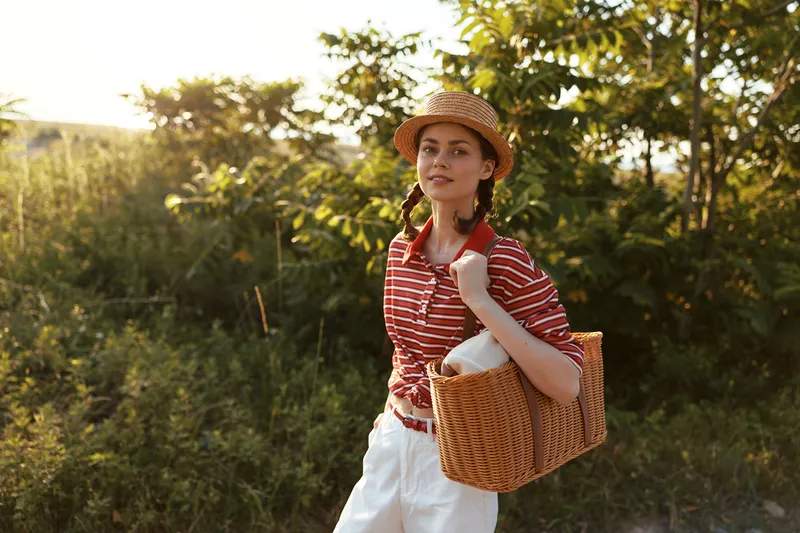 Young woman with a straw hat and a basket standing in a sunny nature park. Lifestyle photo capturing a happy girl enjoying leisure time outdoors during a warm golden hour summer day.