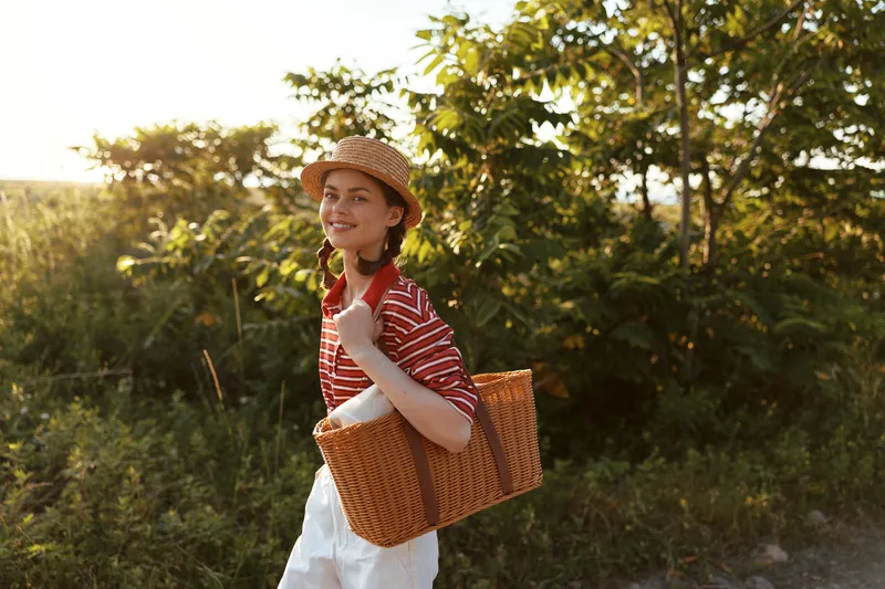 Young woman with braided hair in a straw hat carries a woven picnic basket walking through a sunny garden. Concept for authentic lifestyle leisure and summer relaxation.
