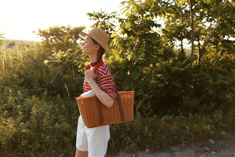 Young woman with braided hair wearing a straw hat walks in a sunny park with a woven basket. The authentic scene captures a peaceful lifestyle moment during a warm summer evening.