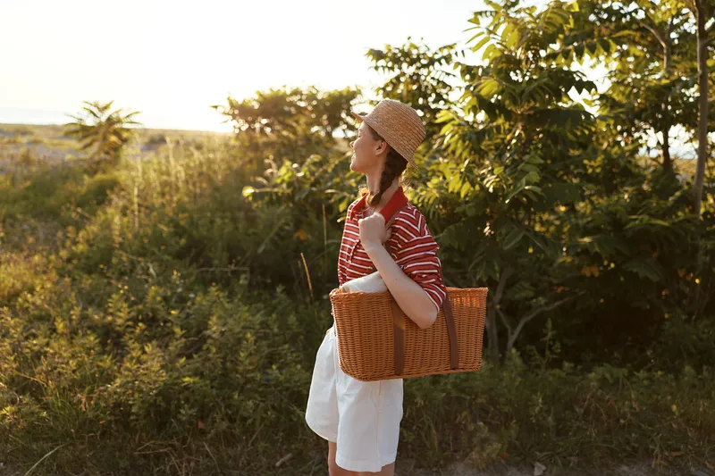 Young woman with basket walking in nature. Lifestyle portrait at sunset. Casual clothing and straw hat in field. Warm golden light creates a peaceful summer mood.