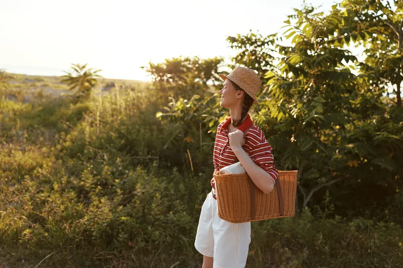Young woman enjoying lifestyle during a summer walk in the park. She carries a straw bag and wears a hat. Sunlight illuminates the nature scene. Concept for vacation, travel, or leisure.