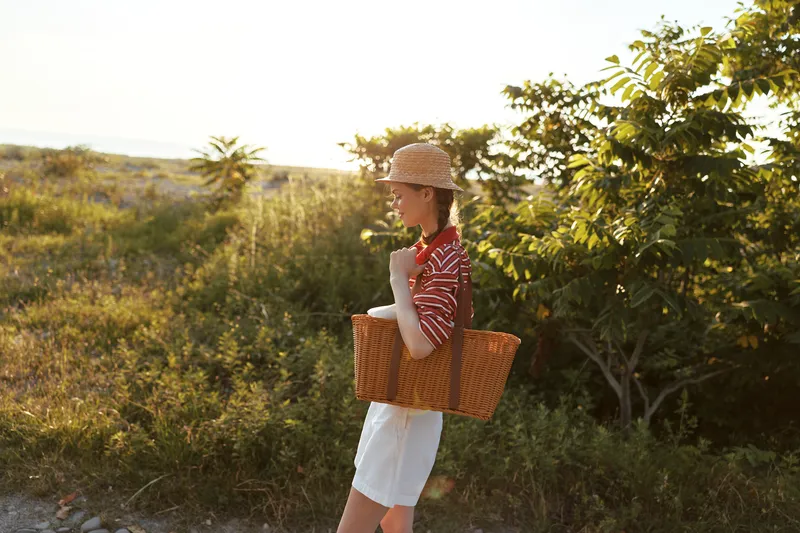 Young woman with a straw hat and wicker basket walks through a sunny meadow during a warm summer evening, embodying a peaceful lifestyle and authentic connection with nature.