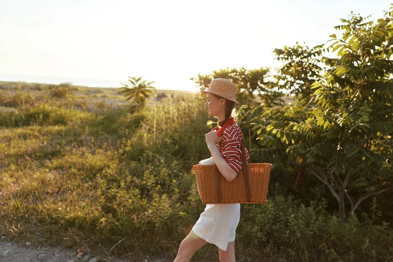 Young woman enjoying a lifestyle walk in the nature carrying a wicker basket. She wears a straw hat and casual summer clothes during golden hour. Concept for weekend leisure and travel trip.