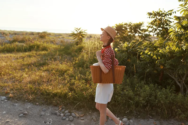 Young woman with basket walking outdoors at sunset, lifestyle portrait. The lady in straw hat enjoys summer nature. Natural soft lighting creates a warm and peaceful atmosphere.