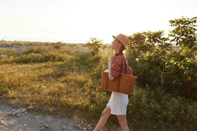 Young woman enjoying lifestyle, walking with a wicker basket in nature during sunset. She wears casual summer clothes and a straw hat. Concept for outdoor leisure.