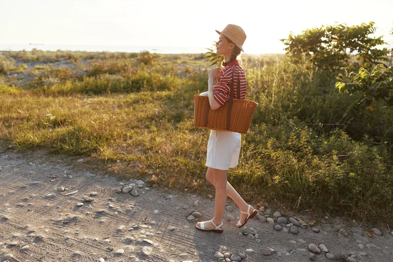 Young woman walking on a countryside road with a picnic basket. She enjoys a summer landscape on a sunny day. Concept for lifestyle, leisure, travel, recreation, outdoor activity, and nature.