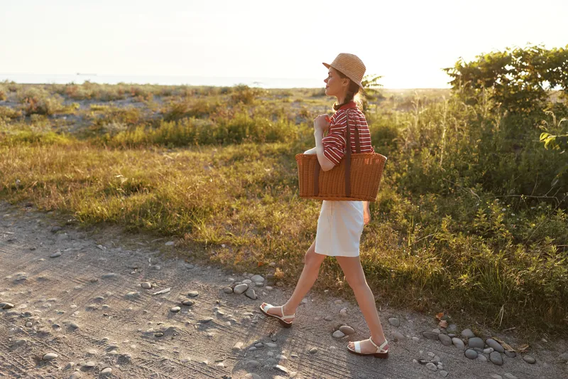 Young woman walking with wicker basket on dirt road during summer sunset. Lifestyle photography capturing travel, vacation, recreation, and peaceful leisure time walk.