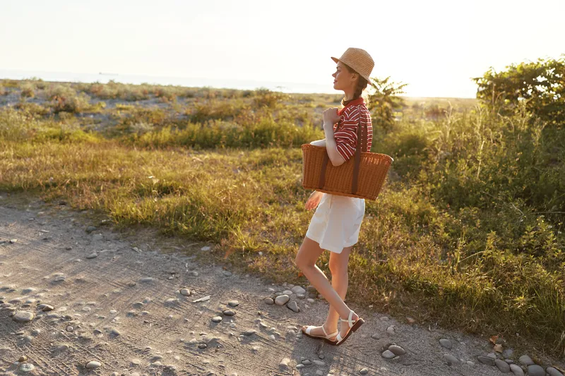 A young woman lifestyle portrait walking on a countryside path with a woven basket. Summer vacation concept with a straw hat and casual outfit in the golden sunset light.