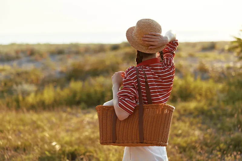 A young woman wearing a straw hat carries a wicker basket in a field. Lifestyle portrait showing a girl enjoying nature during sunset on a summer day. Concept for freedom and rest.