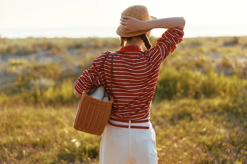 Young woman wearing a straw hat and striped shirt walks through a sunny field carrying a woven basket. This aesthetic lifestyle scene captures a peaceful summer evening mood.