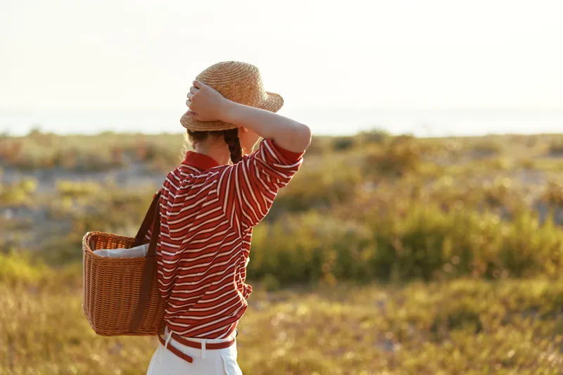Young woman with a straw hat and striped shirt carries a wicker basket while walking through a sunny field during a peaceful summer afternoon, embodying a classic relaxed lifestyle aesthetic.