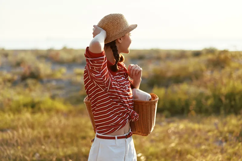 Young woman in a straw hat and striped shirt carries a picnic basket through a golden sunlit meadow enjoying a peaceful summer day in a simple casual style.