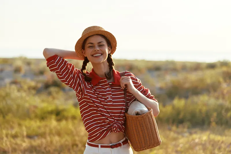 Smiling woman lifestyle portrait wearing a straw hat and striped shirt outdoors. The girl carries a wicker bag while walking in a field. Concept of summer travel, freedom, joy and vacation.