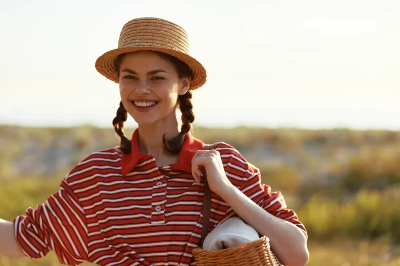 Young woman in a straw hat and striped shirt smiles warmly while carrying a picnic basket outdoors. Authentic lifestyle portrait capturing a cheerful female during a sunny summer vacation day.
