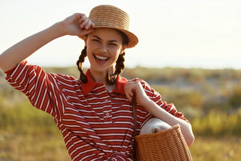 Smiling young woman in a straw hat and striped shirt carrying a wicker basket outdoors. Lifestyle portrait of a happy female enjoying a summer day in nature. Concept of leisure and travel.