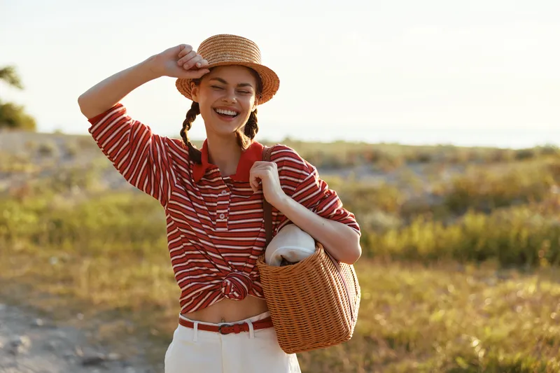 Happy woman lifestyle outdoor portrait, wearing a straw hat and striped shirt, carrying a wicker bag, smiling and enjoying a summer sunny day at the seaside during golden hour.