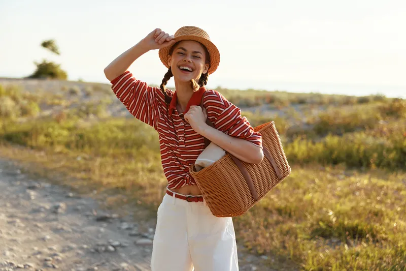 Young woman with pigtails wearing a striped polo and straw hat carries a woven bag while walking outdoor during a sunny summer day. She smiles warmly at the camera with a joyful expression.