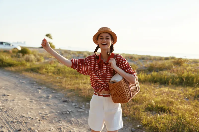 A happy young woman in a hat hitchhiking on a country road holding a basket. Lifestyle portrait of a cheerful female traveler enjoying summer vacations outdoors.