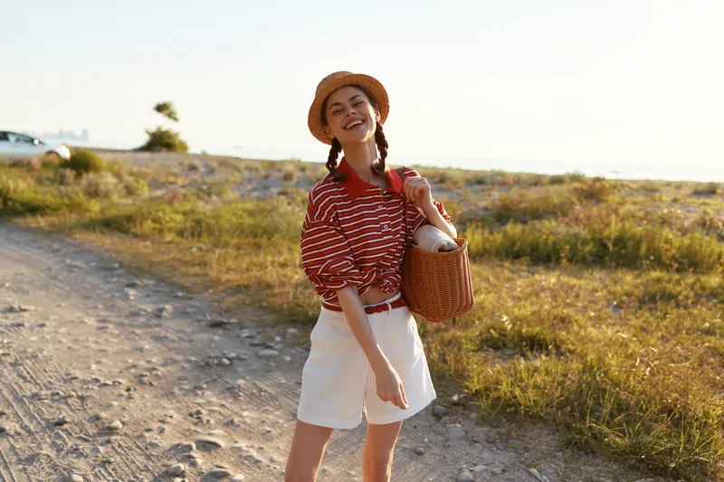 Happy woman enjoying lifestyle during a summer walk on a sandy beach. She wears a straw hat and striped shirt, holding a bag. Sunny outdoor scene with warm lighting.