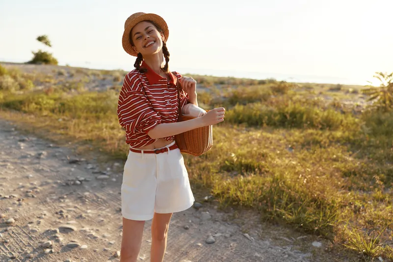 Smiling woman with a basket walks outdoors. Young female lifestyle at sunset in summer. Portrait of a happy lady in a hat enjoying leisure time in nature. Soft light.