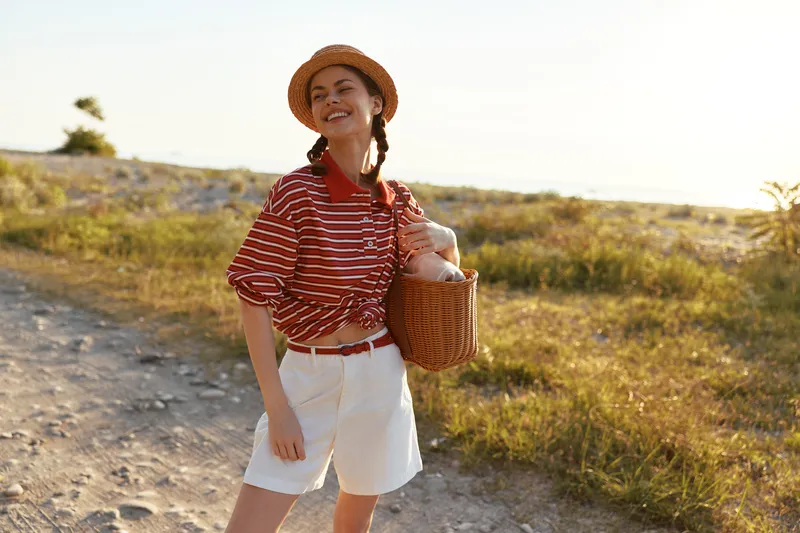 Smiling woman walking outdoors in casual clothes with a straw hat and basket. Lifestyle concept of vacation, summer holidays, travel, and leisure time in nature. Soft golden sunlight at sunset.