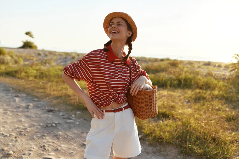 A happy young woman in a straw hat and striped shirt walks along a sunny beach with a basket, embodying a summer lifestyle. Concept for carefree vacation, female joy, and outdoor recreation.