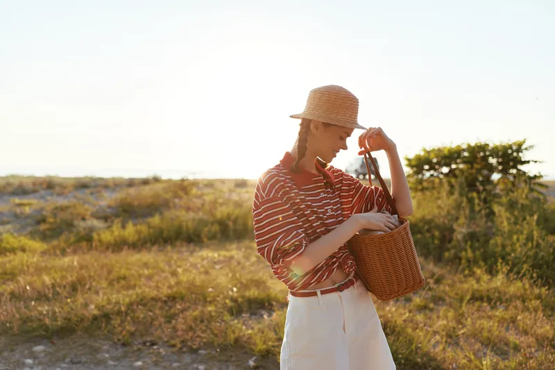 Young woman enjoying lifestyle on a sunny summer day at the beach field. She wears a straw hat and holds a woven basket bag in nature. Concept of vacations, travel, freedom, and happiness.