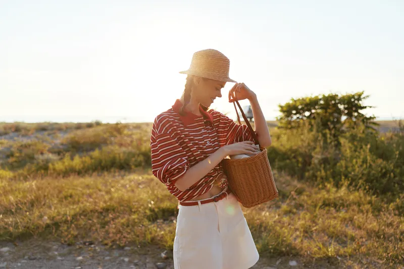 A young woman wearing a straw hat and casual clothes holding a wicker basket during sunset. Lifestyle outdoor concept featuring female style in a bright field for seasonal vacation activity.
