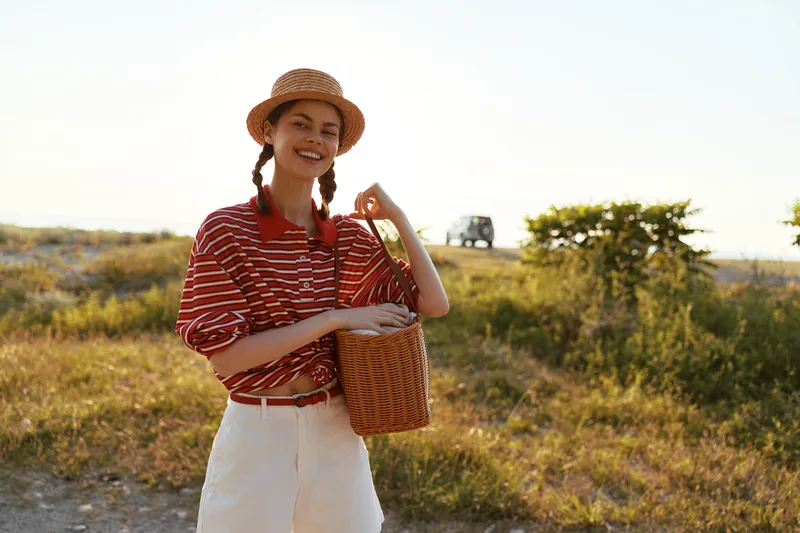 Happy woman lifestyle portrait outdoor in nature wearing striped shirt and straw hat with bag. Authentic young female traveler enjoying sunny day during summer vacation. Natural golden light.