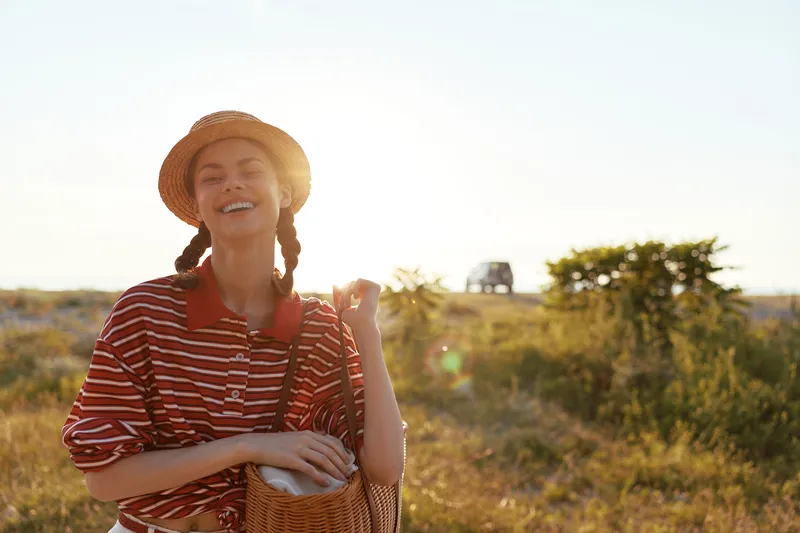 Young woman in a straw hat and striped shirt smiles while walking outdoors during a sunny summer vacation trip. Authentic lifestyle portrait with warm natural light.