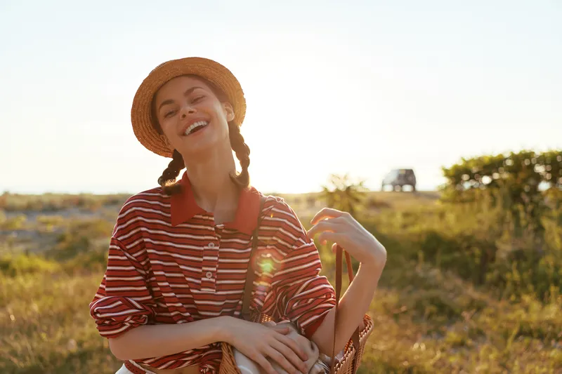 A joyful young woman wearing a straw hat and a striped shirt laughs during a sunny summer vacation in a field at sunset, capturing an authentic lifestyle moment of freedom and happiness.